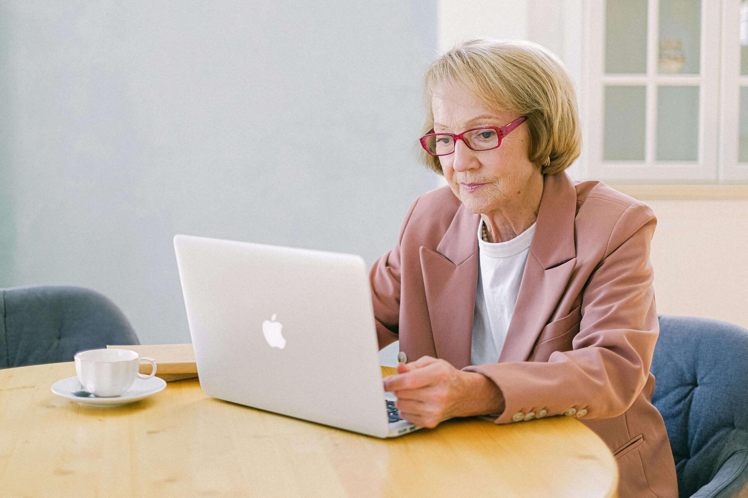 Elderly woman using a laptop for online grief therapy with a cup of coffee on the table.