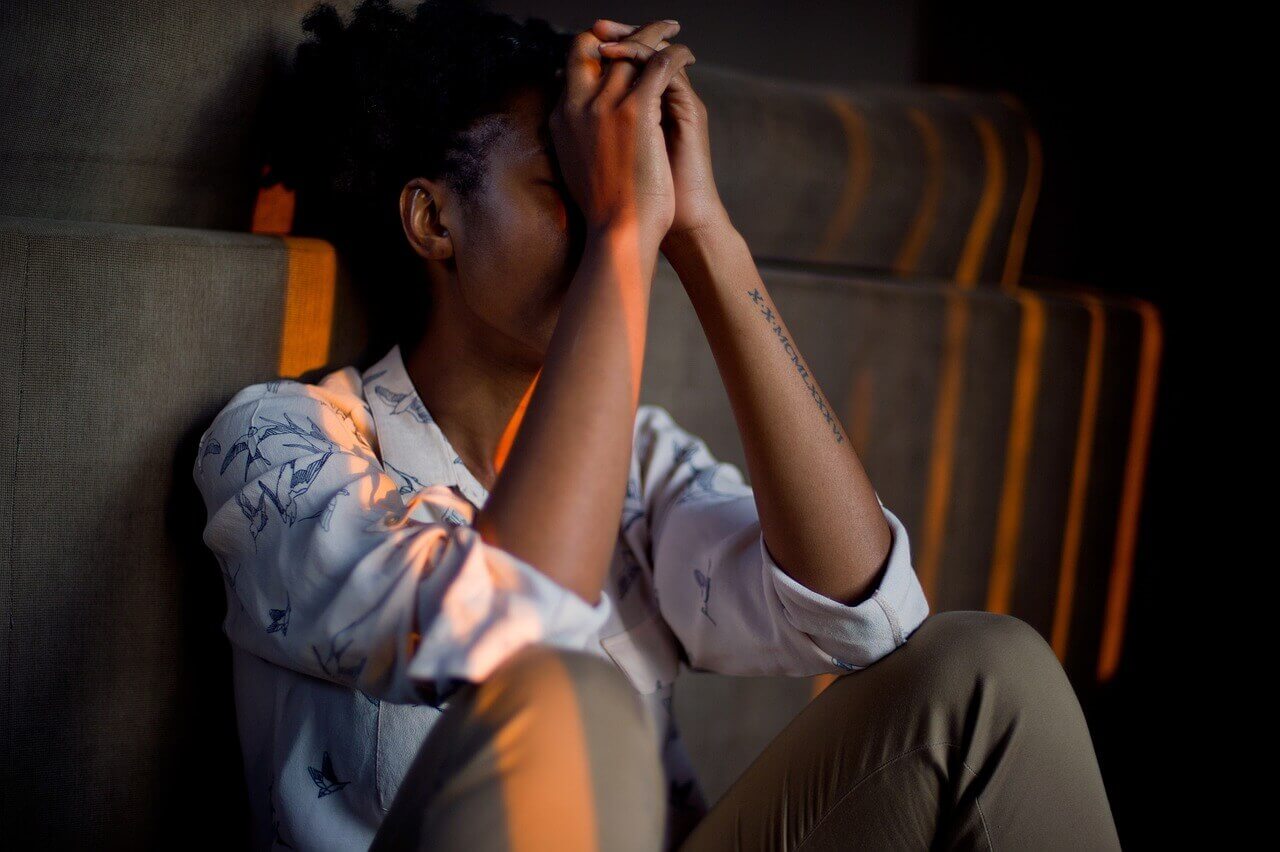 Person sitting against a wall with hands clasped, wearing a patterned shirt, in a contemplative pose.