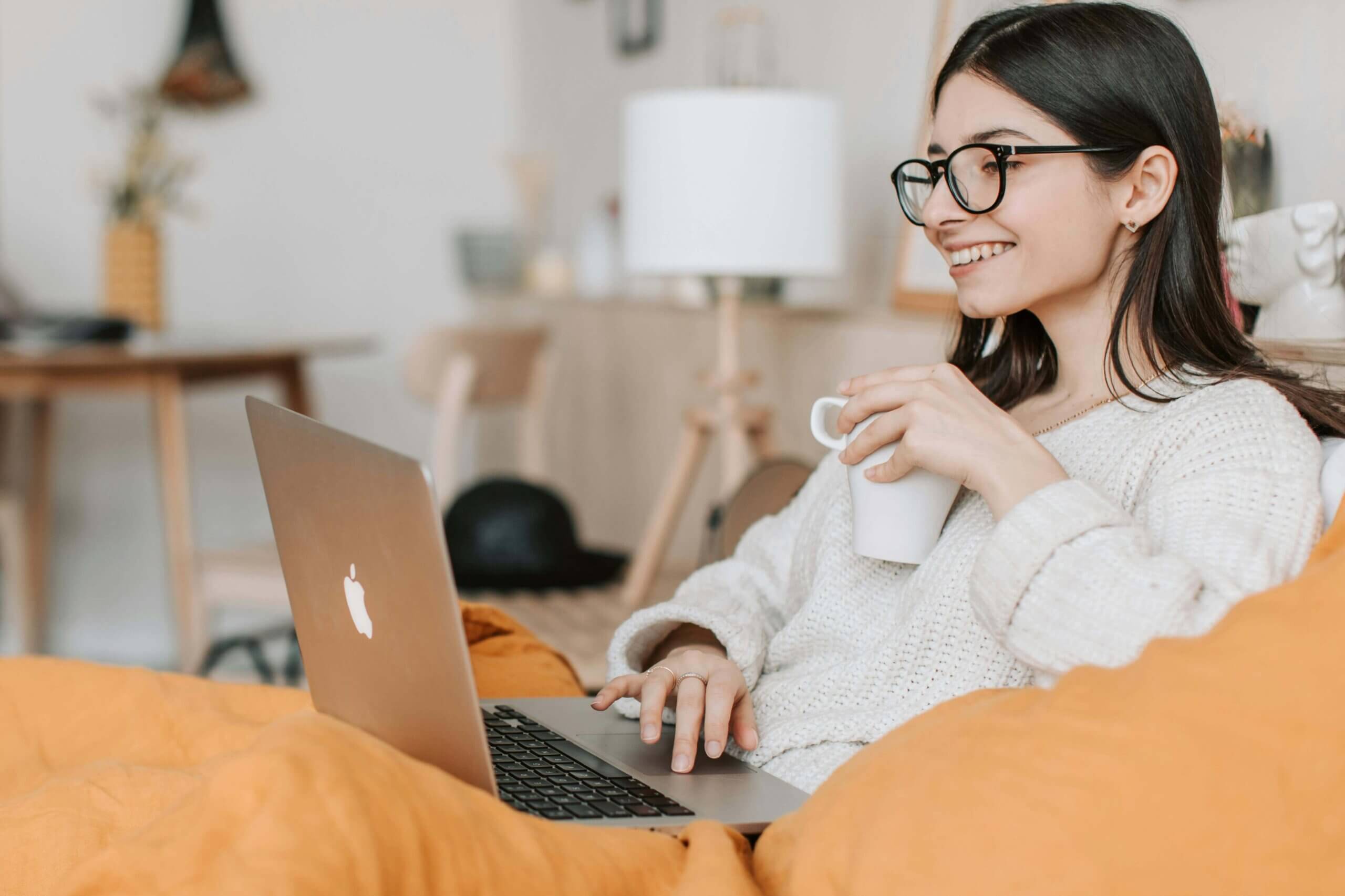 Woman using a MacBook while drinking coffee, representing online telehealth psychiatry in a cozy home setting.